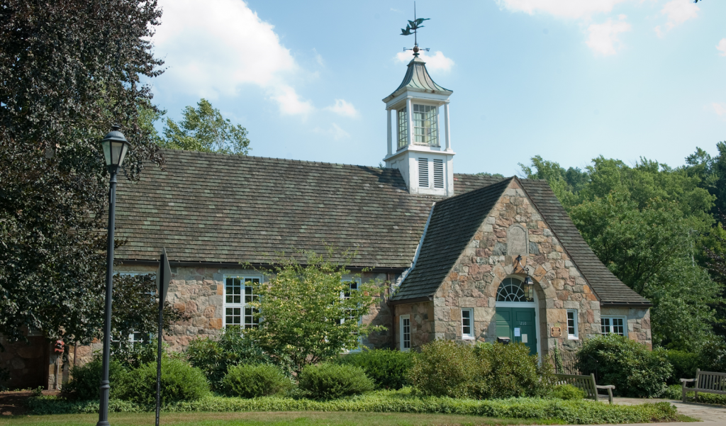 a historic multi-colored fieldstone building with a rustic, cottage-like aesthetic. A prominent gabled entrance on the right with a dark green arched door and a small lantern above it. A white wooden cupola sits atop the roof, topped with a weather vane featuring a bird.