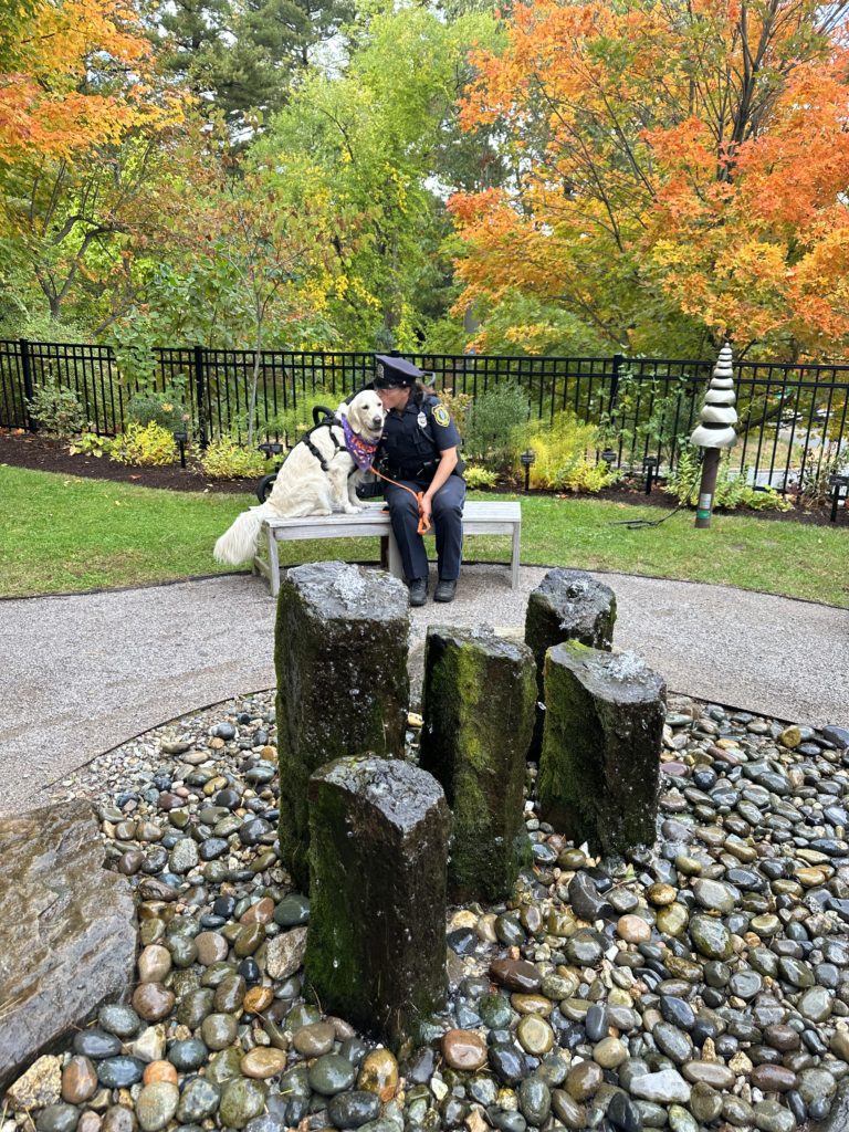 A photo of a police officer sitting with an emotional support dog in front of the fountain in the garden.