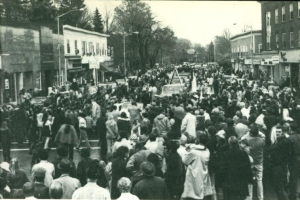 A black and white, high-angle photograph showing a large crowd of people packed onto a town main street during an event. Several vintage cars and a vehicle with a triangular structure on top are visible amidst the sea of people. The street is lined with multi-story commercial buildings and bare trees