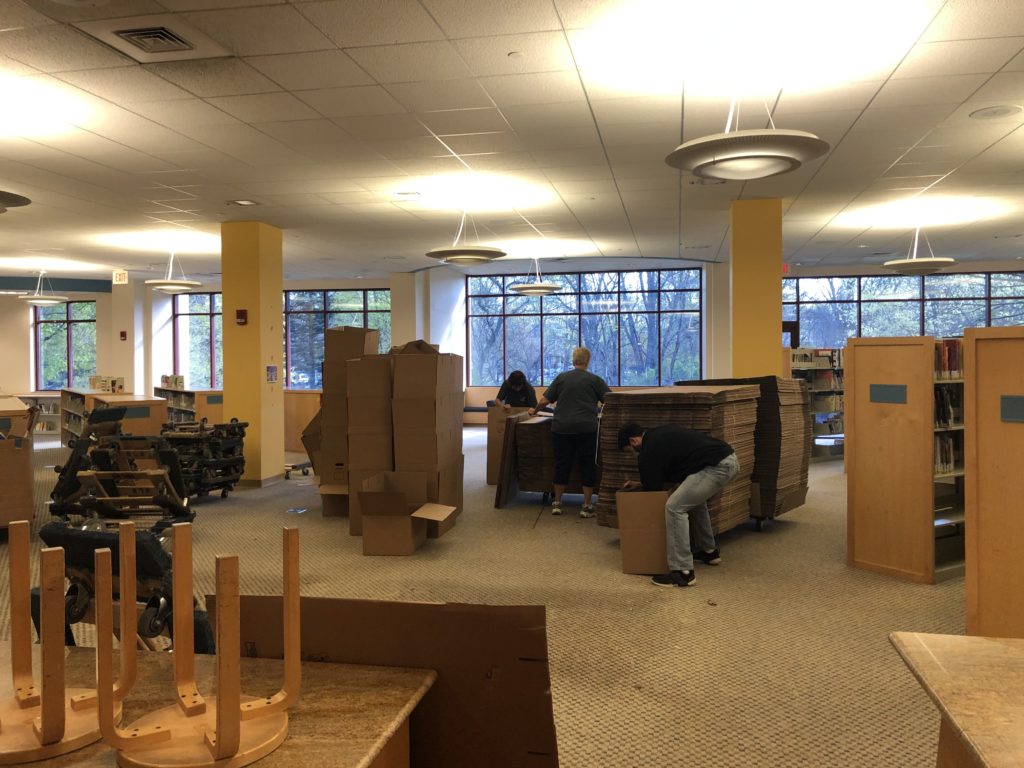 Three workers bring dollies to the old children's room piled high with boxes to be assembled. There's a stack of assembled boxes that's taller than one of the workers and another pile of dollies next to that.