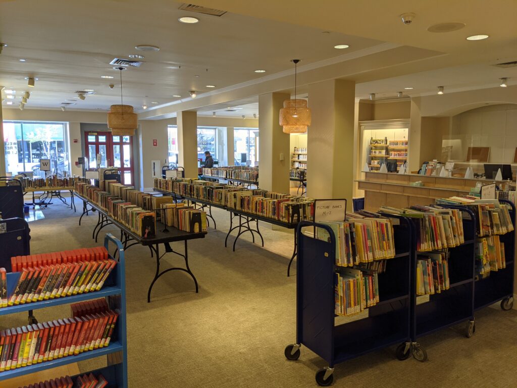 Adult books on tables, playaways, and picture books on book carts, and in the back, books on CD lining the shelves. Another checkout counter is repurposed into the circulation desk.