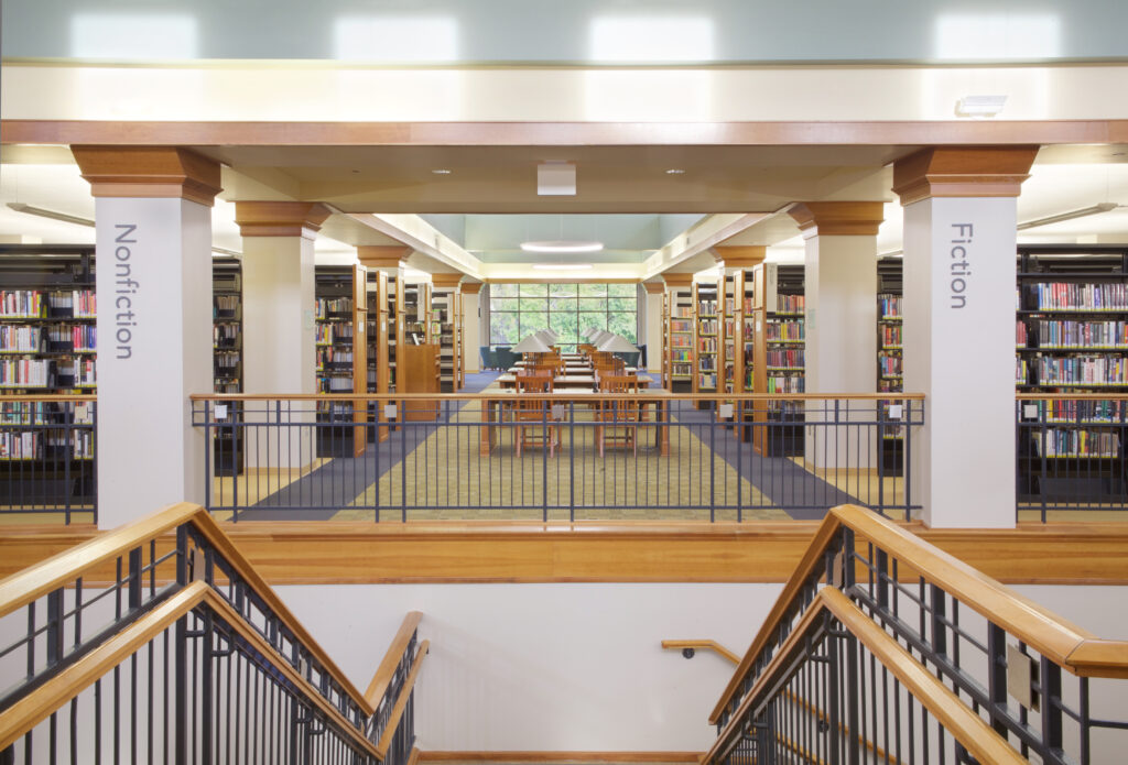 The second floor library stacks, as viewed from the Information desk. The collection is split into Nonfiction on the left and Fiction on the right and is labeled with signs on the closest pillars.