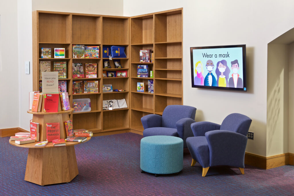 The Welcome Center seating area, next to shelving for the Library of Things collection. A display on this wall is used to promote library events and P.S.A.s.