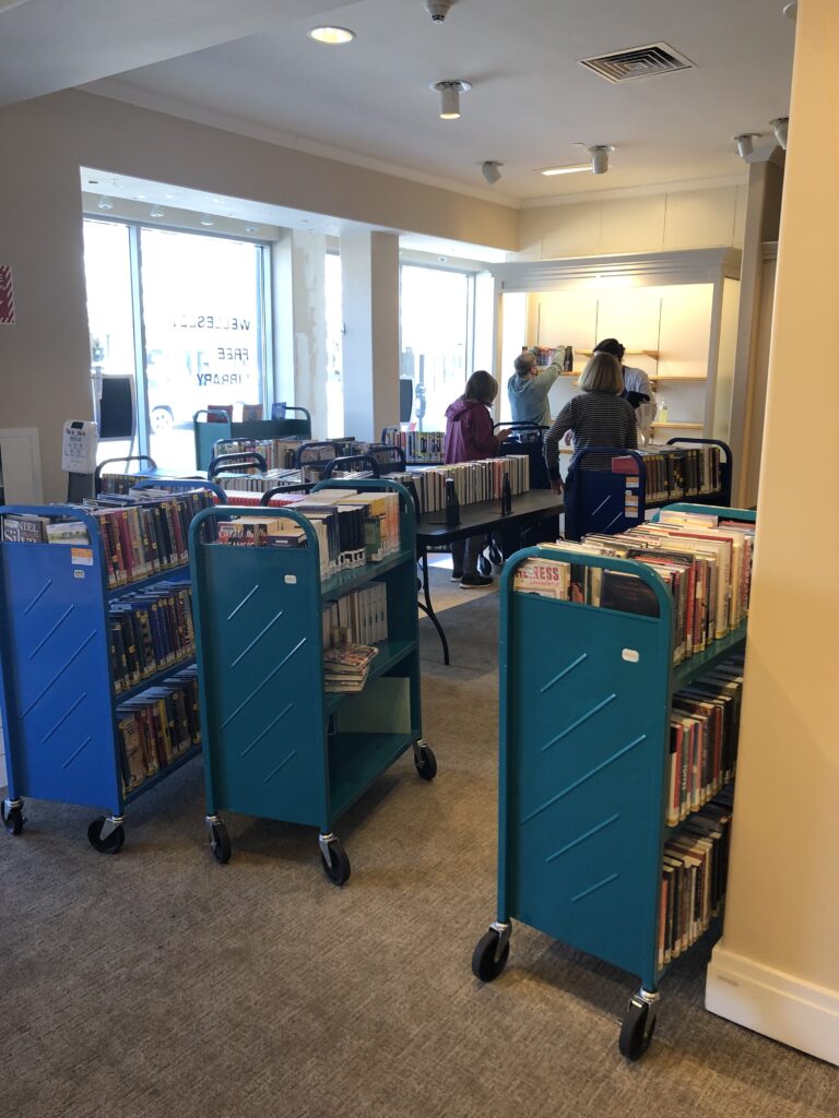 Book carts with new items, summer reading books, and some general collection books lined up to help populate the space. Librarians are preparing the shelves and adding items to folding tables for display. The words Wellesley Free Library can faintly be seen decorating the window.