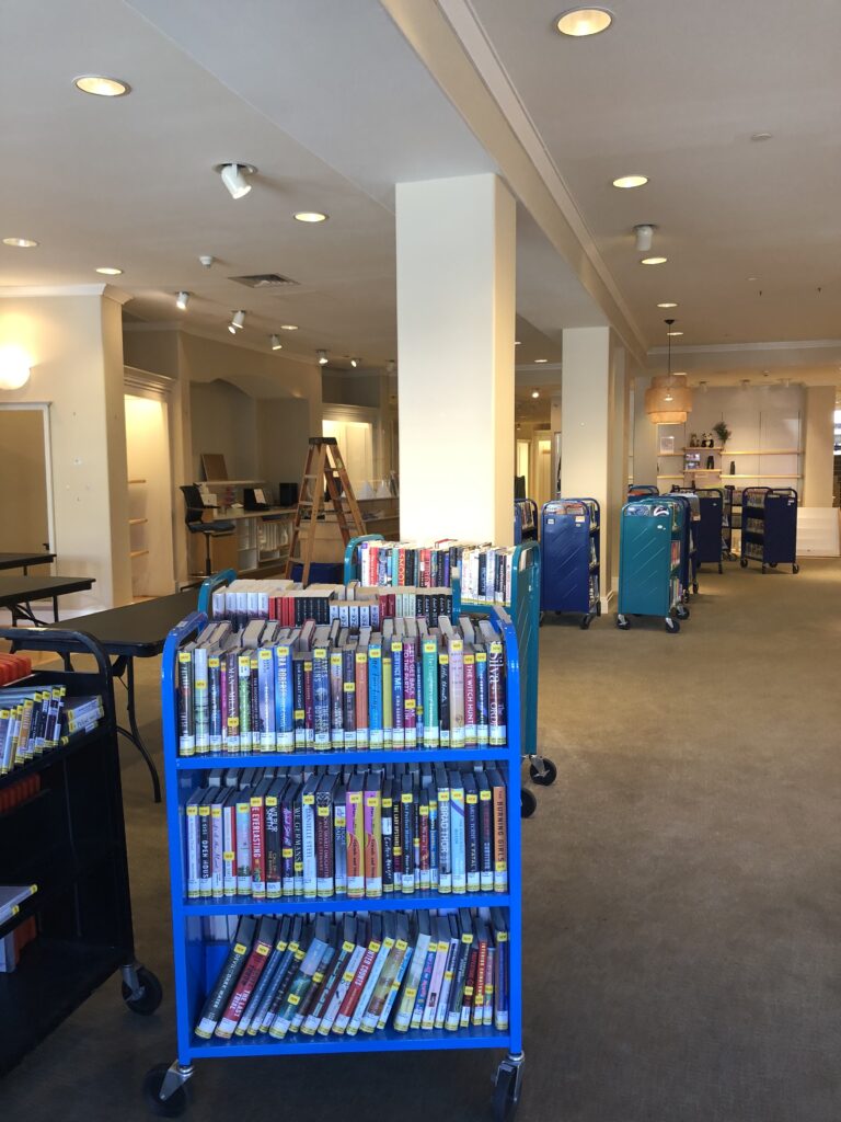 Full book carts scattered around, amongst more folding tables. An old check out desk in the background has been repurposed into a makeshift workstation.