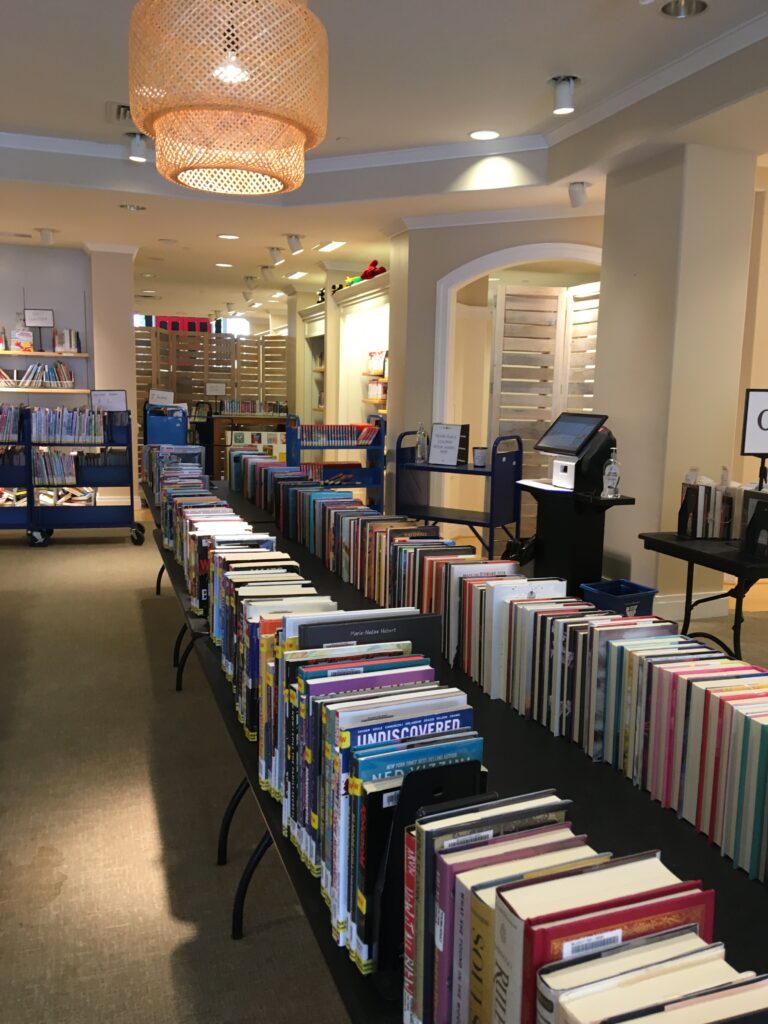 Rows of books displayed on folding tables. There is a self-check in the background, right in front of the old dressing room. The space is much more brightly lit and welcoming.