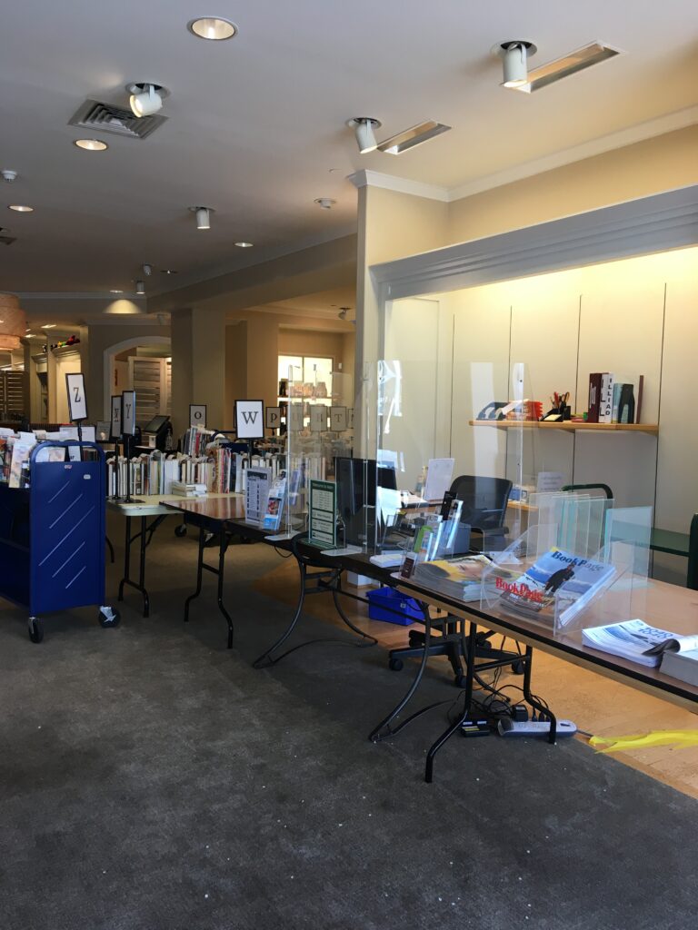 The makeshift Information Services desks set up on folding tables. Book carts with holds on them are beyond the desks. Magazines, tax prep forms, and the Librarian's binder for information on Inter-Library Loan can also be seen.