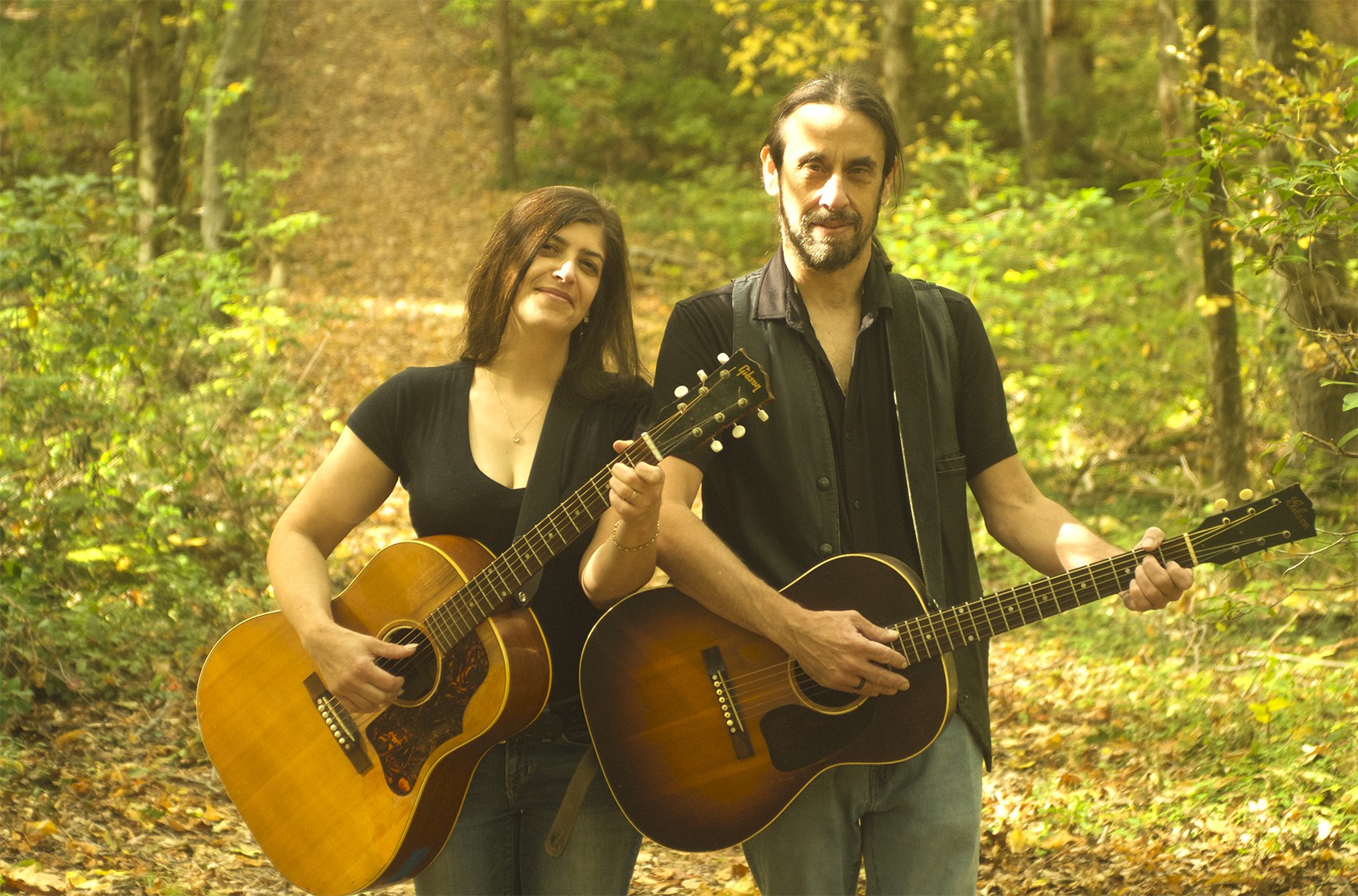 A man and woman standing in the woods, with acoustic guitars, for an upcoming Wellesley Free Library program.
