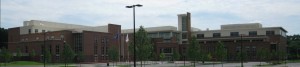 Wide view of a large modern brick-and-glass building with a central tower, trees in front, and parking lot lights in the foreground.